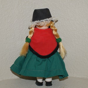 May include: A porcelain doll wearing a traditional folk costume. The doll has blonde braids, a black hat with white lace trim, a red bodice, and a green skirt.