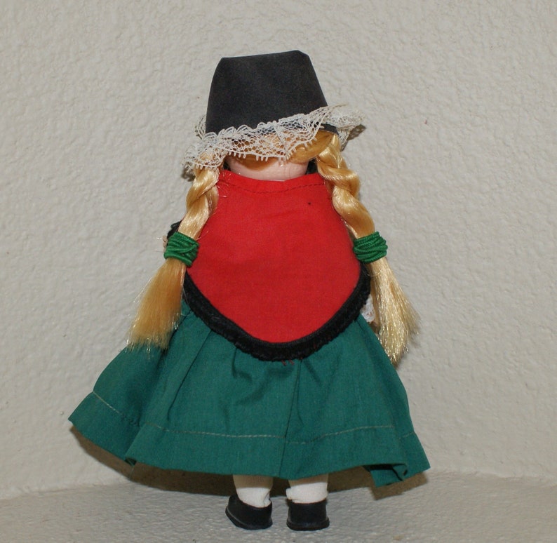 May include: A porcelain doll wearing a traditional folk costume. The doll has blonde braids, a black hat with white lace trim, a red bodice, and a green skirt.