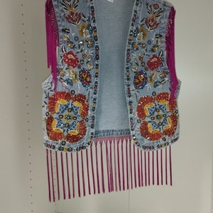 Op de afbeelding: Een denim vest met geborduurde bloemenmotieven en roze franjes. Het vest heeft een maatlabel met de tekst "M".