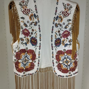 Op de afbeelding: Witte, mouwloze vest met gouden franjes en kleurrijke bloemenborduurwerk. De vest is versierd met kralen en pailletten.