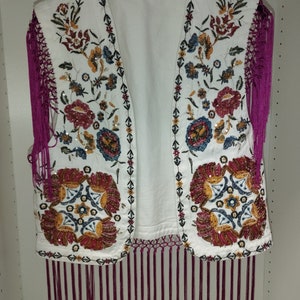 Op de afbeelding: Een wit vest zonder mouwen met een geborduurd bloemenmotief en een franje rand. De borduurwerk is versierd met gekleurde kralen en pailletten.