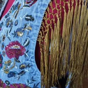Op de afbeelding: Een lichtblauw denim vest met geborduurde bloemenmotieven en gouden franjes. Het vest is mouwloos en heeft een V-hals.
