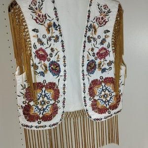 Op de afbeelding: Witte, mouwloze vest met gouden franjes en kleurrijke bloemenborduursel. Het borduursel is versierd met kralen en pailletten.