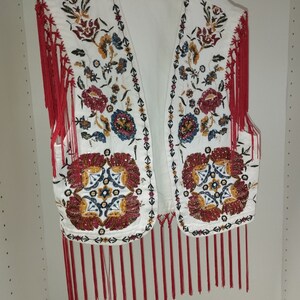 Op de afbeelding: Witte, mouwloze vest met rode franjes en kleurrijke bloemenborduursels. De vest is versierd met kralen en pailletten.