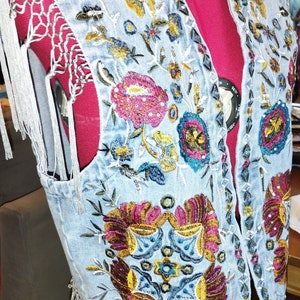 May include: A light blue denim vest with colorful floral embroidery. The embroidery features a variety of flowers, leaves, and vines in shades of pink, yellow, blue, and green. The vest has a fringe trim around the edges.