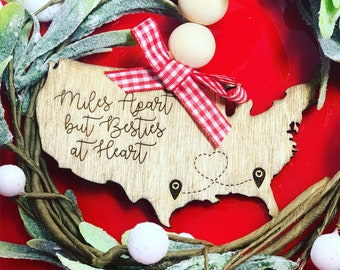 May include: Wooden ornament shaped like the United States with the text "Miles Apart but Besties at Heart" and a red and white gingham bow. The ornament has two small heart-shaped pins with a dotted line connecting them.