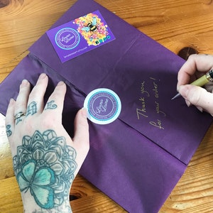 Op de afbeelding: Paarse verpakking met een handgeschreven notitie met de tekst "Bedankt voor uw bestelling!" en twee ronde stickers met de tekst "Dream and Painted, Lorna Laine" en een bij-illustratie.