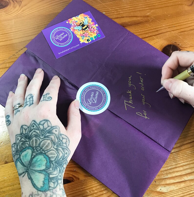 Op de afbeelding: Paarse verpakking met een handgeschreven notitie met de tekst "Bedankt voor uw bestelling!" en twee ronde stickers met de tekst "Dream and Painted, Lorna Laine" en een bij-illustratie.