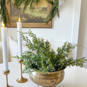 May include: A hammered brass bowl filled with artificial green cedar branches and frosted blue berries, accompanied by two white taper candles in tall brass candlesticks with glowing yellow LED flames, creating a warm and festive display.
