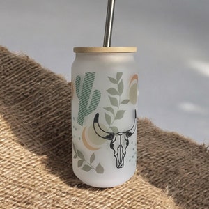 May include: A white frosted glass tumbler with a bamboo lid and a stainless steel straw. The tumbler has a desert-themed design with a cactus, leaves, and a skull with horns.
