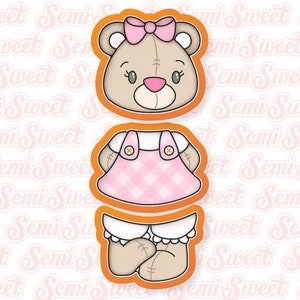 May include: A three-piece cookie cutter set featuring a teddy bear wearing a pink and white dress. The bear has a pink bow on its head and brown eyes.