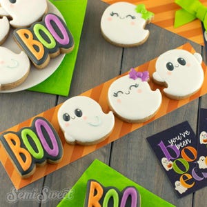 May include: A collection of Halloween-themed sugar cookies featuring white ghosts with black eyes and pink cheeks, some wearing purple or green bows, and cookies shaped like the word BOO in orange, green, and purple icing on a dark background.