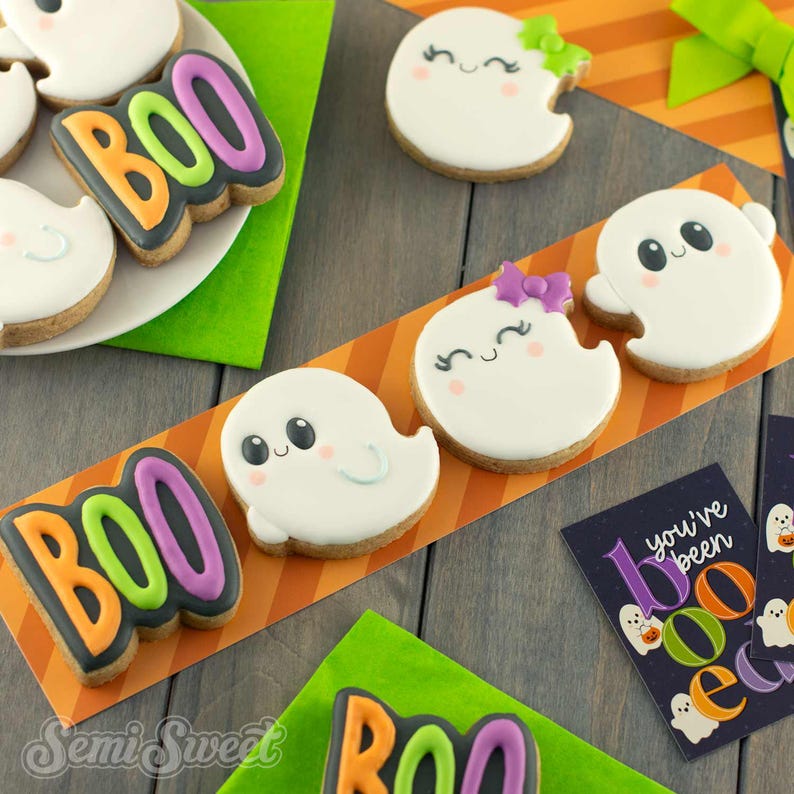 May include: A collection of Halloween-themed sugar cookies featuring white ghosts with black eyes and pink cheeks, some wearing purple or green bows, and cookies shaped like the word BOO in orange, green, and purple icing on a dark background.