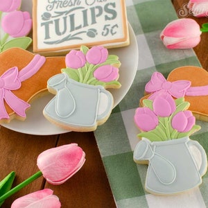 May include: A collection of decorated sugar cookies, including a hat with a pink bow, a watering can with pink tulips, and a sign that reads "Fresh Tulips 5&cent;". The cookies are arranged on a white plate and a green and white checkered cloth.
