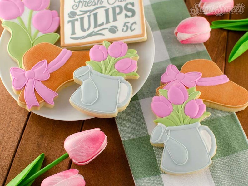 May include: A collection of decorated sugar cookies, including a hat with a pink bow, a watering can with pink tulips, and a sign that reads "Fresh Tulips 5&cent;". The cookies are arranged on a white plate and a green and white checkered cloth.