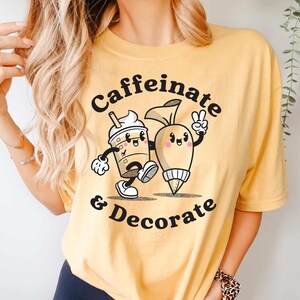 Pode incluir: Uma camiseta amarela com um gr&aacute;fico de uma x&iacute;cara de caf&eacute; e um saco de confeitar com o texto "Caffeinate & Decorate".