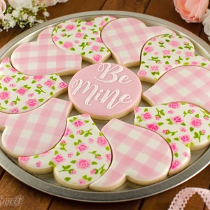 Peut inclure: Une assiette de biscuits en forme de cœur rose et blanc décorés de motifs vichy et floraux. Le biscuit du centre dit "Be Mine".
