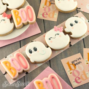 May include: A collection of Halloween-themed sugar cookies featuring white ghost shapes with black eyes and pink cheeks, some wearing pink bows, alongside cookies shaped like the word BOO in orange and pink icing, with matching printed cards.