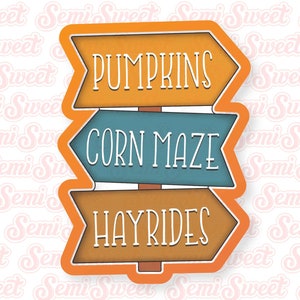 May include: A brown, blue, and orange wooden sign with three arrows pointing in different directions. The arrows have the words "Pumpkins", "Corn Maze", and "Hayrides" written on them.