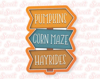 May include: A brown, blue, and orange wooden sign with three arrows pointing in different directions. The arrows have the words "Pumpkins", "Corn Maze", and "Hayrides" written on them.