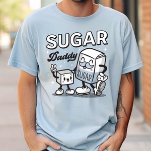 May include: A light blue t-shirt with a cartoon illustration of two sugar cubes. The larger sugar cube has a mustache and says "Sugar Daddy" and "Sugar" on it. The smaller sugar cube has a happy face.