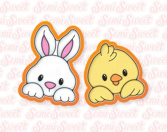 May include: Two cookie cutters, one shaped like a bunny rabbit and the other shaped like a chick. Both are in an orange outline and have a white background.