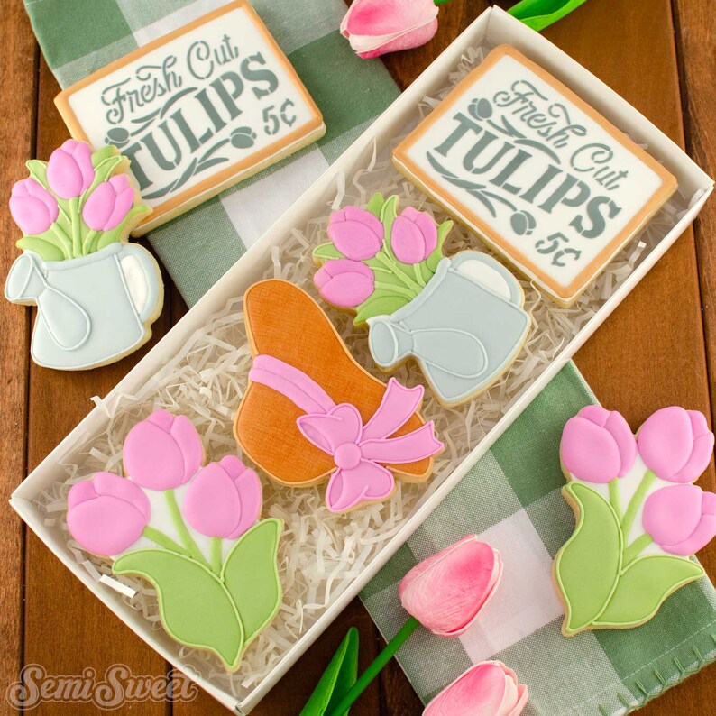 May include: A white box filled with decorated sugar cookies. The cookies are shaped like tulips, watering cans, a sun hat with a pink bow, and signs that read "Fresh Cut Tulips 5&cent;". The cookies are decorated with pink, green, and light blue icing.