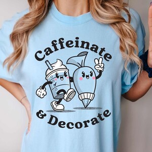 Pode incluir: Uma camiseta azul claro com um gr&aacute;fico de uma x&iacute;cara de caf&eacute; de desenho animado e um saco de confeitar com o texto "caffeinate & decorate".