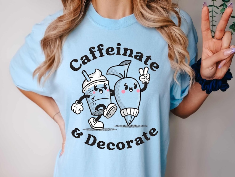 Pode incluir: Uma camiseta azul claro com um gr&aacute;fico de uma x&iacute;cara de caf&eacute; de desenho animado e um saco de confeitar com o texto "caffeinate & decorate".