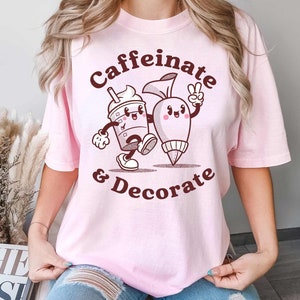 Pode incluir: Camiseta rosa com um gr&aacute;fico marrom de uma x&iacute;cara de caf&eacute; e um saco de confeitar com o texto "Caffeinate & Decorate".
