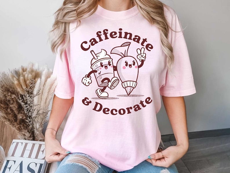 Pode incluir: Camiseta rosa com um gr&aacute;fico marrom de uma x&iacute;cara de caf&eacute; e um saco de confeitar com o texto "Caffeinate & Decorate".