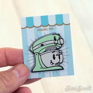 May include: A mint green enamel pin shaped like a stand mixer with a smiling face on the mixer head and the mixing bowl. The pin is attached to a light blue and white striped backing card that features the text ENAMEL PIN at the top.
