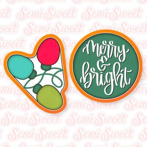 May include: Two cookie cutters, one shaped like a circle with the text "Merry & Bright" in white lettering on a green background, and the other shaped like a string of Christmas lights with red, green, and blue bulbs.