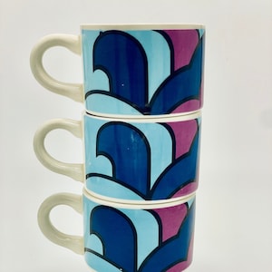May include: A set of three white ceramic mugs with blue and pink abstract geometric patterns. The mugs are stacked on top of each other.