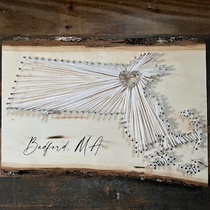 May include: Wooden string art of the state of Massachusetts, with a heart-shaped design. The word "Bedford, MA" is written in cursive below the state outline. The artwork is mounted on a light-colored wooden plank with a natural edge.