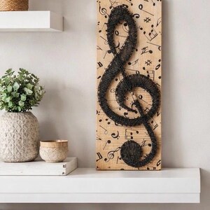 May include: A wooden wall art piece featuring a black treble clef made of wire. The background is a light brown wood with musical notes printed on it. The artwork is displayed on a white shelf with decorative items.