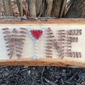 May include: Wooden sign with the word "WINE" spelled out in a string art design. The letters are made with brown string and nails, with a red wine glass in the center. The sign is on a light wood background.