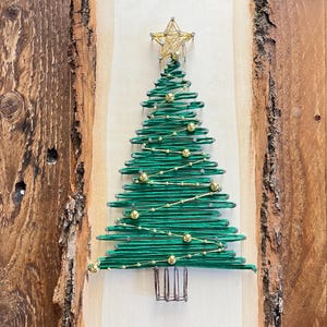 May include: A handmade Christmas tree string art piece. The green tree is crafted with string on a light wood background, adorned with gold beads and a gold star topper. The wood is mounted on a rustic, bark-edged wooden board.