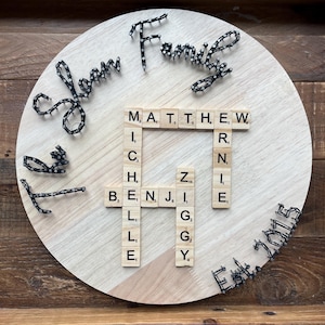 Può includere: Un cartello di famiglia rotondo in legno con le parole "John Family" ed "Est. 2015" in filo nero. I nomi dei membri della famiglia sono scritti con tessere di legno del gioco dello Scarabeo. Il cartello è presentato su un vassoio di legno.