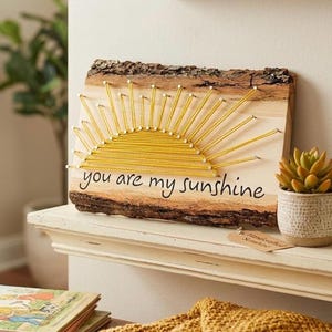 May include: A rectangular wooden plaque with natural bark edges featuring a yellow string art sun design. The text "you are my sunshine" is printed in black cursive lettering below the sun. The piece is crafted with nails and yellow thread.
