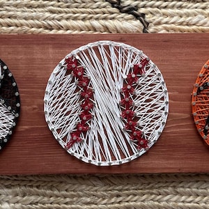 May include: A wooden plaque featuring string art representations of a soccer ball, baseball, and basketball. The soccer ball is black and white, the baseball is white with red accents, and the basketball is orange with black details.