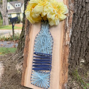 May include: A wooden wall hanging featuring a string art design of a vase filled with yellow flowers. The vase is crafted with light blue string, and the base has dark blue horizontal lines. The flowers are a bright yellow.