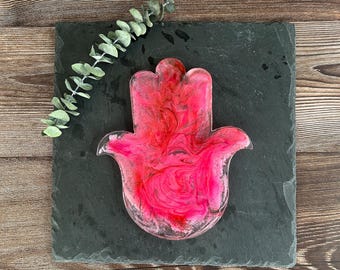 May include: A pink and red resin Hamsa hand, a symbol of protection, sits on a dark gray square slate. The resin has a swirled pattern. A sprig of green eucalyptus is placed on the slate. The background is a wooden surface.