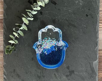 May include: A blue resin Hamsa hand-shaped coaster with glitter accents. The coaster has a clear border and is filled with shades of blue and silver glitter. The coaster is displayed on a dark gray slate surface.