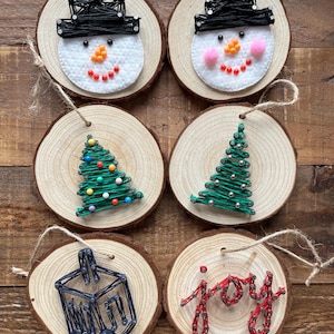 May include: Six handmade wooden Christmas ornaments. Two snowman decorations with black hats and orange and red details. Two green Christmas tree decorations with colourful and silver accents. One dreidel and one ornament that says "joy" in red.