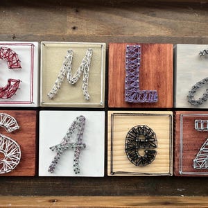 May include: A collection of eight wooden blocks, each featuring a letter of the alphabet crafted with string art. The letters are in various colors, including red, silver, purple, and black, set against different wood tones and white backgrounds.