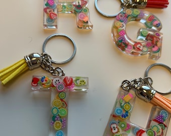 May include: Four clear resin keychains shaped as the letters R, G, T, and H. Each letter is filled with colorful fruit-shaped polymer clay pieces. Each keychain has a silver ring and a tassel in green, red, yellow, and orange.