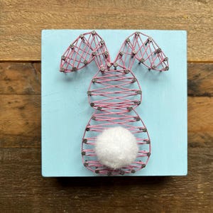 May include: A string art bunny design on a light blue square wooden block. The bunny is crafted with pink string and silver-colored nails, featuring a fluffy white pom-pom tail. The background is a rustic wooden surface.