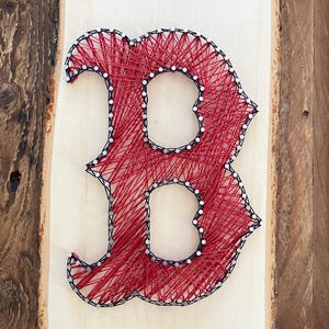 May include: A handmade string art piece featuring the letter "B" in red string, with a black outline, on a light-colored wooden board. The nails are silver. The wooden board has a natural edge and is mounted on a wooden background.