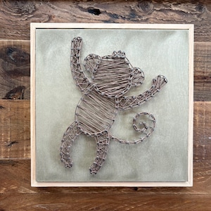 May include: A handcrafted wire monkey art piece. The monkey is depicted in a playful pose, with its arms raised and a curled tail. The artwork is set against a light green background within a wooden frame.